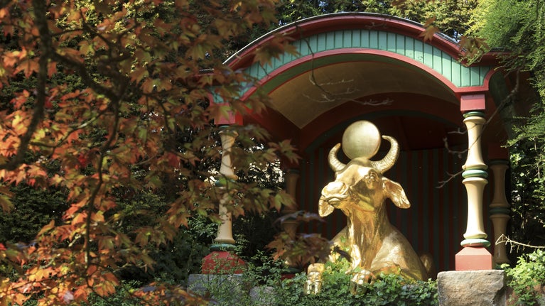The Golden Water Buffalo in the area known as 'China' at Biddulph, Staffordshire. The buffalo is seen lying down with its head up and a sun disc between its horns. The statue is surrounded by trees turning red and brown in autumn.